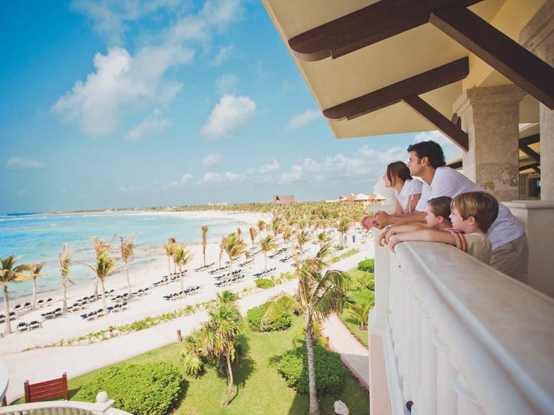 Famiglia guarda da un balcone una spiaggia soleggiata con palme e lettini.