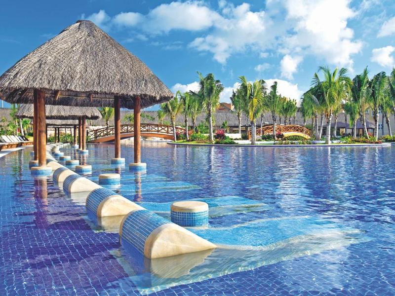 Grande piscina con sgabelli da bar in acqua sotto tetto di paglia e palme sullo sfondo.