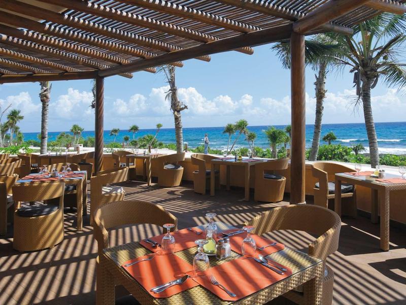 Terrazza coperta con tavoli e sedie con vista sul mare e palme.