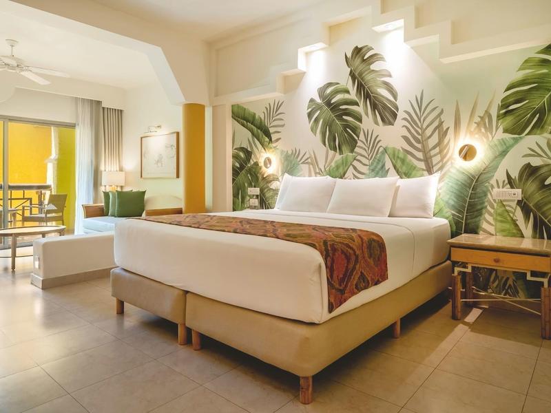 Camera d'hotel moderna con letto grande, decorazione murale tropicale e balcone con area salotto.