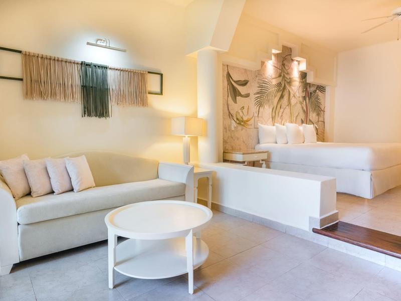 Camera d'hotel luminosa con divano bianco, tavolo rotondo e letto con decorazioni da parete.