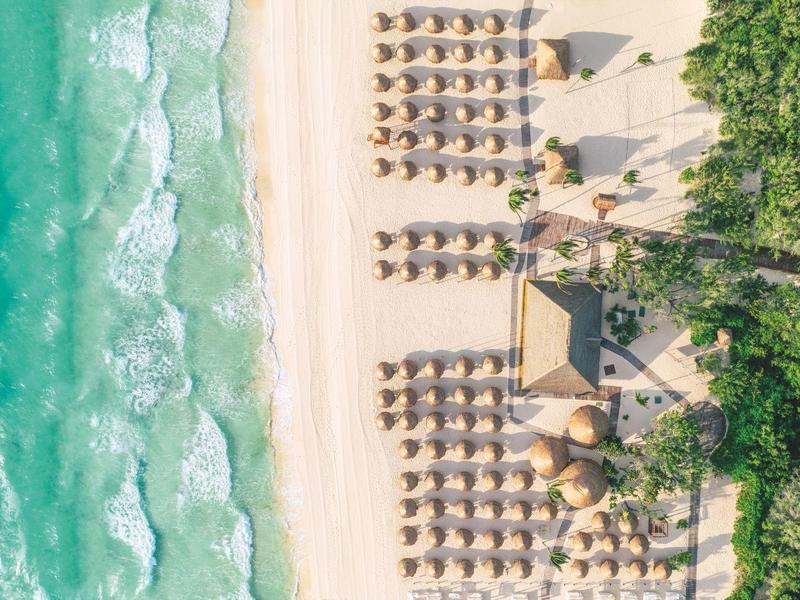 Veduta aerea di una spiaggia con ombrelloni e acqua turchese limpida accanto a una zona boschiva.