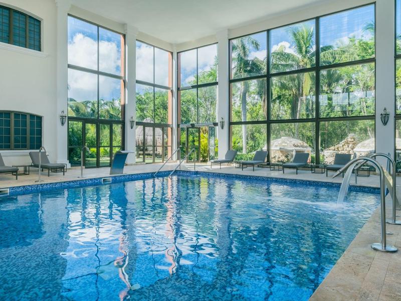 Piscina interna con grandi finestre e lettini, circondata da paesaggio verde.