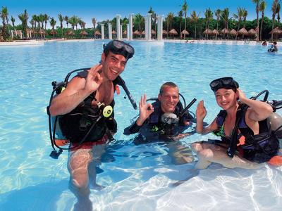 Tres personas en una piscina con equipo de buceo, rodeadas de palmeras y cielo azul.
