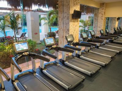 Equipamiento moderno de gimnasio junto a grandes ventanas con vista a la piscina y palmeras.