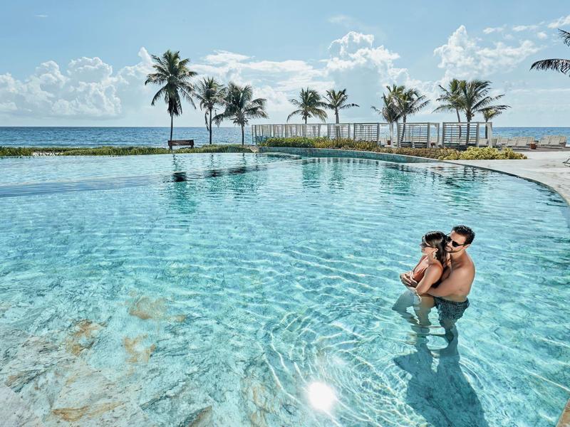 Piscina dell'hotel con vista mare, palme e coppia in acqua.
