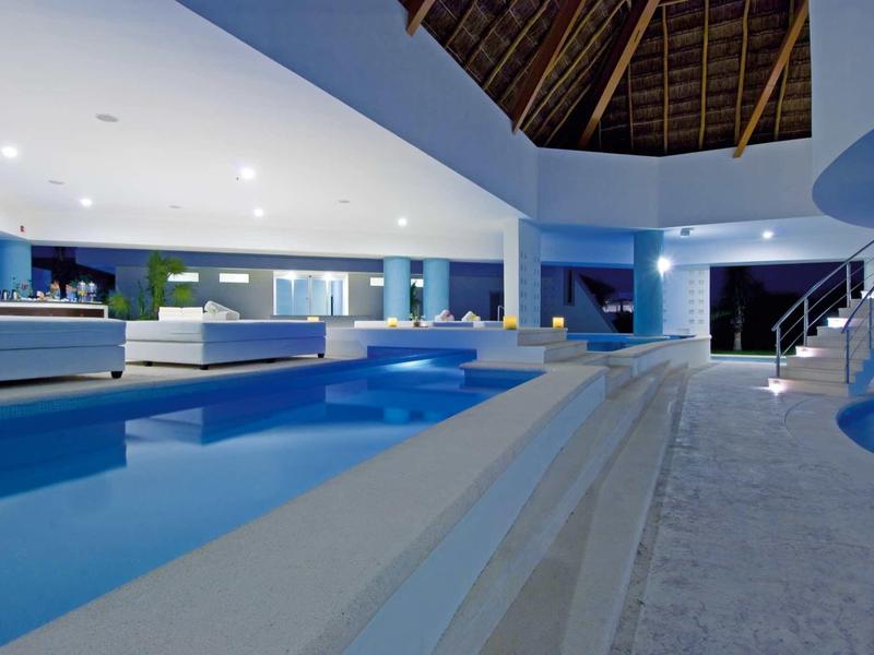 Piscina interna moderna con illuminazione blu e aree lounge sotto un alto soffitto di legno.