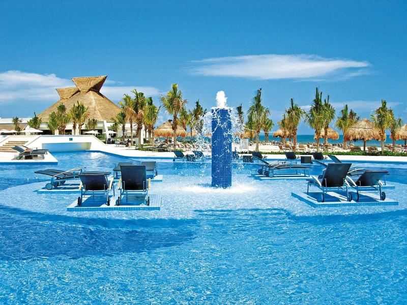Grande piscina con lettini e fontana davanti a palme e cielo limpido.