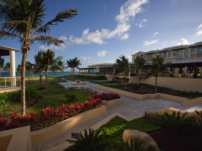 Complesso alberghiero moderno con palme, aiuole e piscina sotto un cielo azzurro al tramonto.