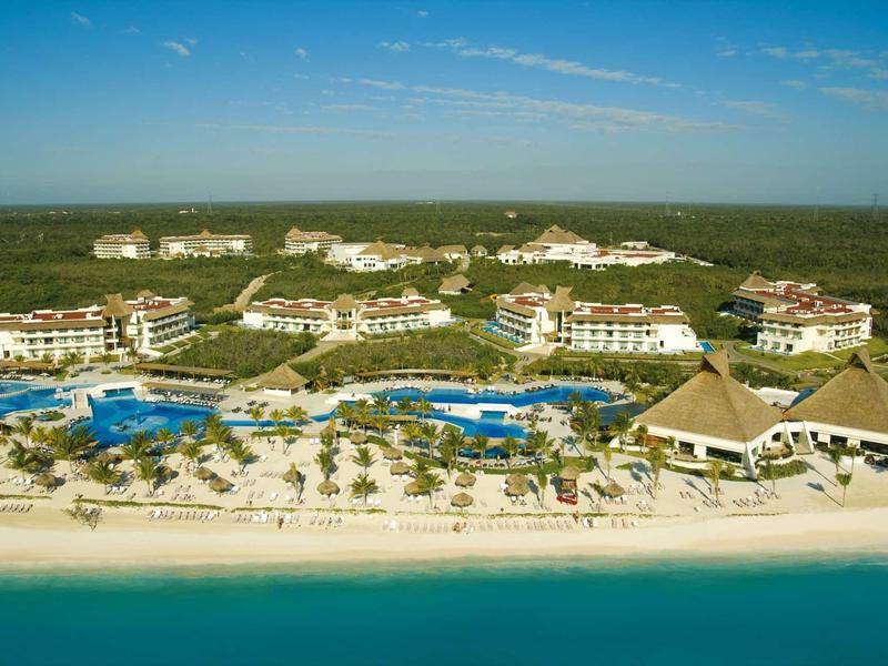 Vista aerea di un villaggio vacanze sulla spiaggia con piscine, bungalow e mare blu sullo sfondo.