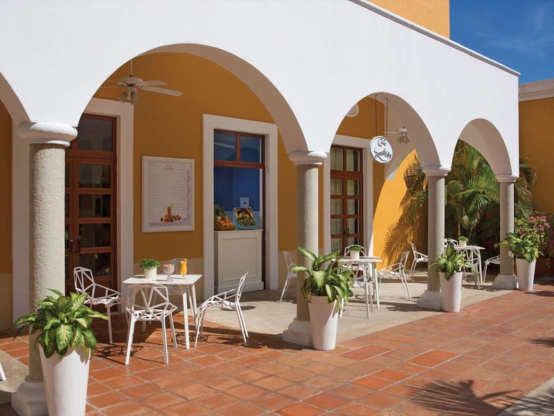 Area esterna con sedie sotto archi bianchi, muri gialli e piante in vaso su pavimento piastrellato.