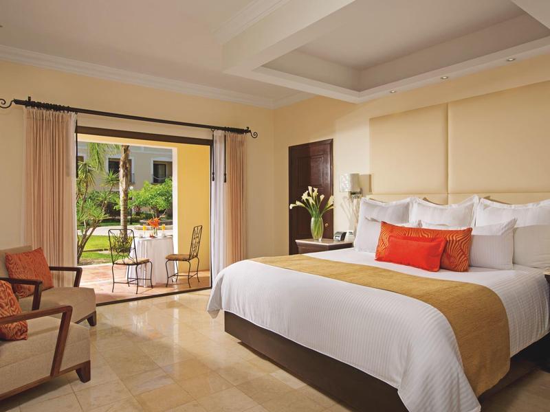 Camera d'hotel luminosa con grande letto, cuscini arancioni, poltrona e balcone con vista giardino.