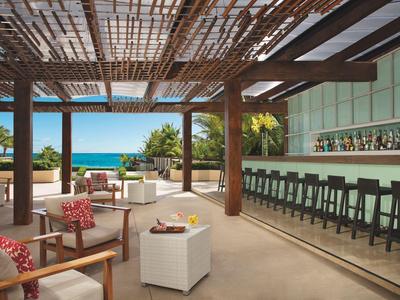 Offene Strandbar mit Holzdach, hoher Bar mit Hockern und bequemen Sitzbereichen mit Blick auf das Meer.