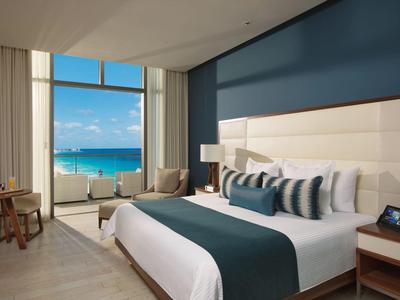 Modern hotel room with large bed, seating area, and ocean view through floor-to-ceiling windows.