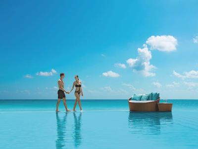 Paar hält Händchen und geht am Rand eines Infinity-Pools am Meer entlang bei klarem Himmel.