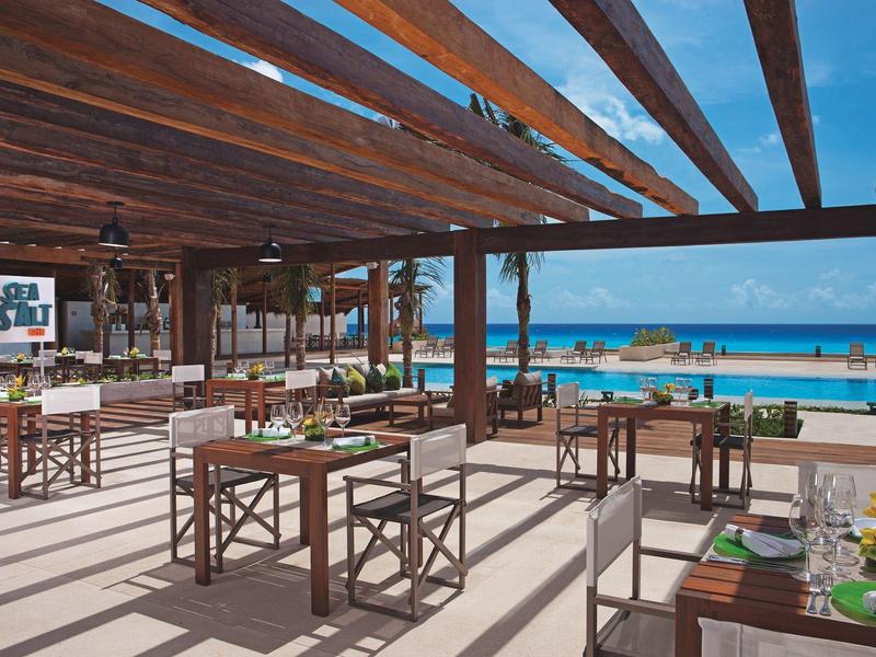 Open restaurant area with wooden furniture overlooking pool and sea under a blue sky.