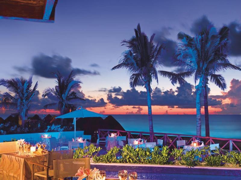 Romantická večeře u bazénu s palmami a výhledem na oceán při západu slunce.