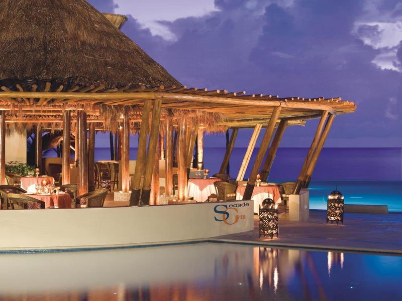 Otevřený bar s doškovou střechou u vody za soumraku s modrou oblohou a klidným mořem.