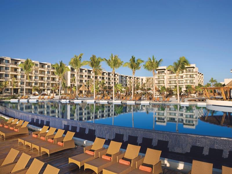 Velký hotelový bazén s lehátky a palmami před vícepodlažní hotelovou budovou za jasné oblohy.