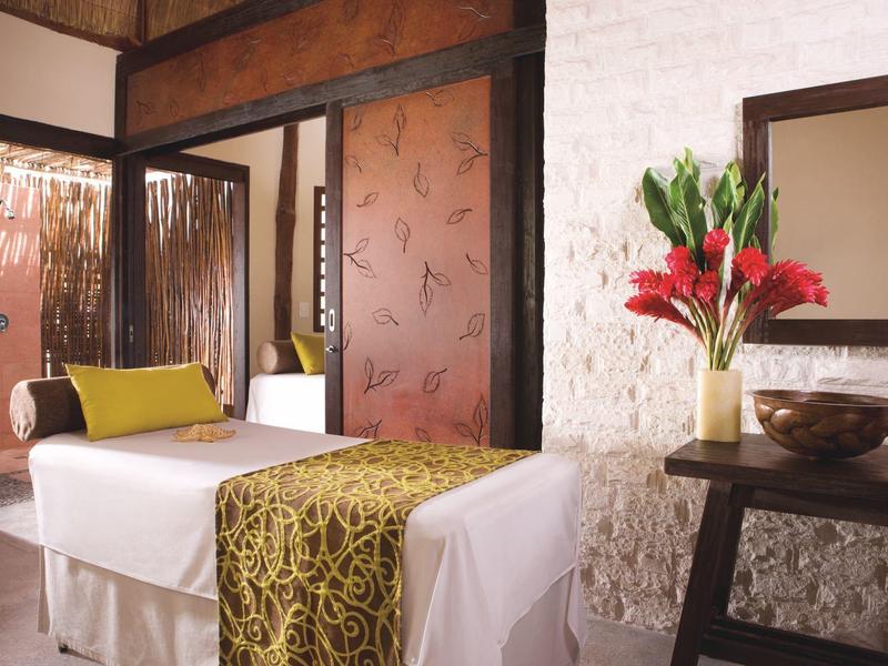 Masážní stůl s bílým potahem a zelenou látkou v klidné místnosti s rostlinou a dřevěnou výzdobou.