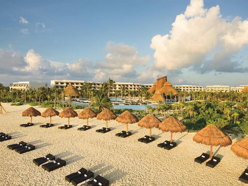 Hotel sulla spiaggia con sdraio e ombrelloni di paglia sotto un cielo blu con nuvole bianche.