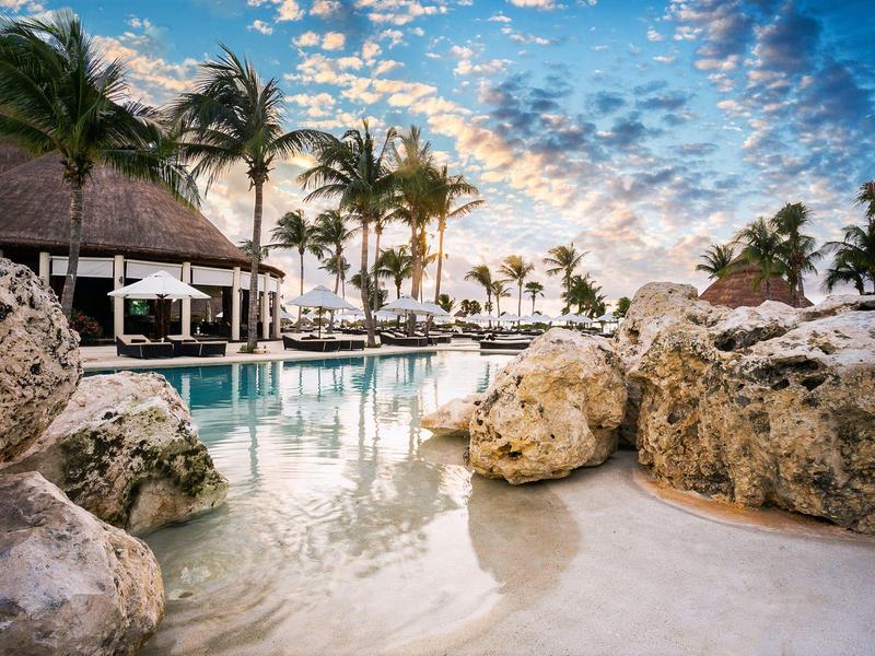 Piscina di hotel di lusso con grandi rocce, palme e un cielo drammatico al tramonto.