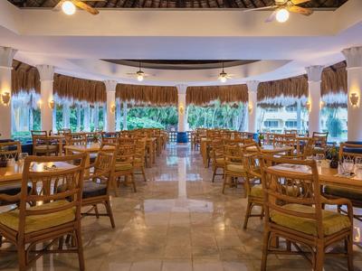 Großer, heller Restaurantraum mit Holzstühlen, Tischen und großen Fenstern mit Vorhängen.