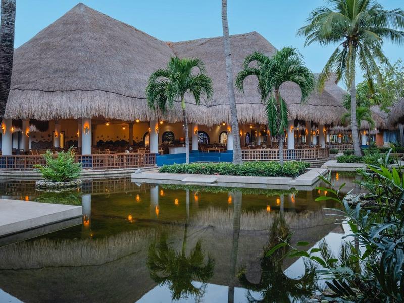 Accogliente resort con tetto di paglia, illuminato, circondato da palme e uno stagno in primo piano.