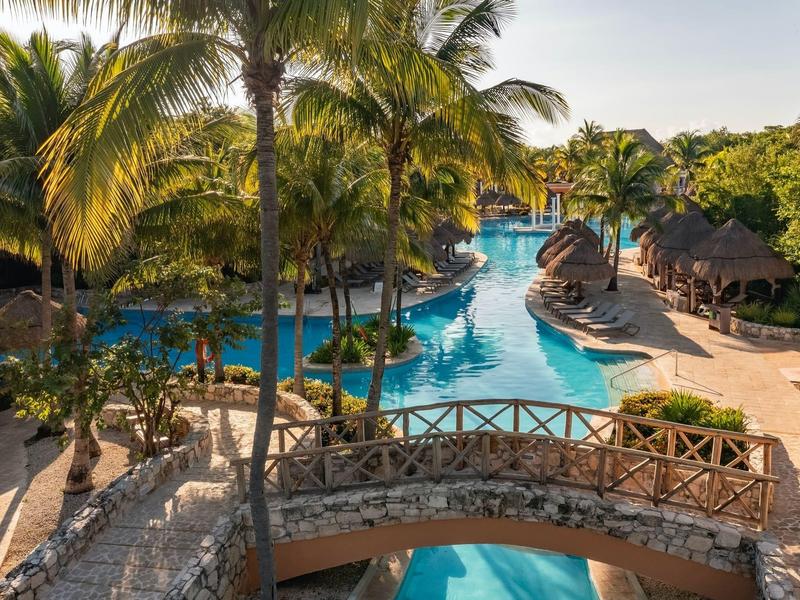 Piscina di hotel di lusso con palme, ponte sospeso e terrazze soleggiate in un ambiente tropicale.