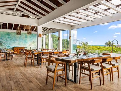 Espace repas ouvert avec tables et chaises en bois, grandes fenêtres donnant sur un paysage vert et un ciel bleu.
