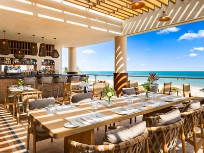 Restaurant ouvert avec des tables en bois offrant une vue sur la plage et la mer sous un ciel bleu.