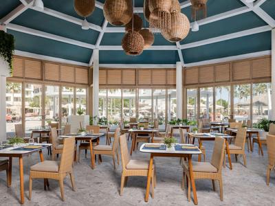 Restaurant lumineux avec tables en bois, chaises et grandes fenêtres dans un cadre hôtelier de vacances.