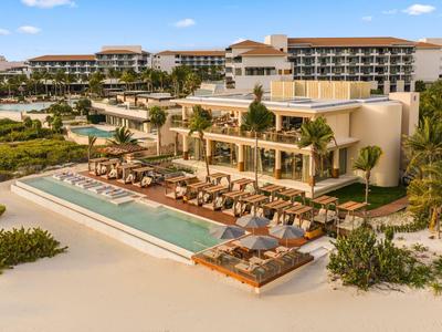 Complex hôtelier de luxe en bord de mer avec piscine, chaises longues et grands bâtiments hôteliers sur une plage de sable blanc.