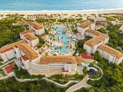 Grand complexe hôtelier avec piscine, palmiers et plage près de l'océan en arrière-plan.