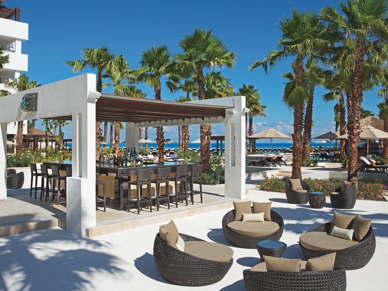 Bar e lounge all'aperto sulla spiaggia con palme e vista oceano sotto cielo sereno.