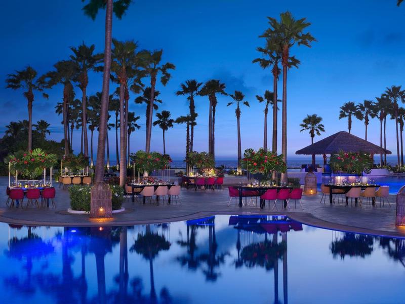 Area piscina esterna al tramonto con palme, lettini e riflessi di luci nell'acqua.