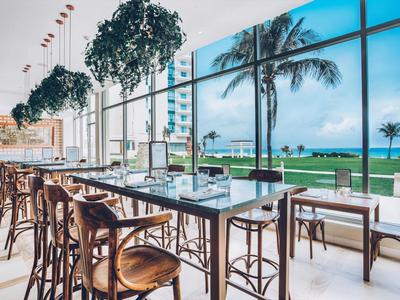 Nowoczesna restauracja z wysokim sufitem, roślinami i widokiem na palmy i morze przez duże okna.