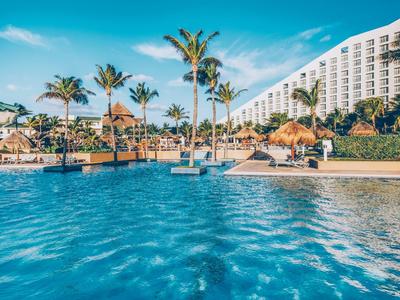 Großer blauer Pool mit Palmen, Sonnenschirmen und modernem Hotelgebäude im Hintergrund bei Sonnenschein.