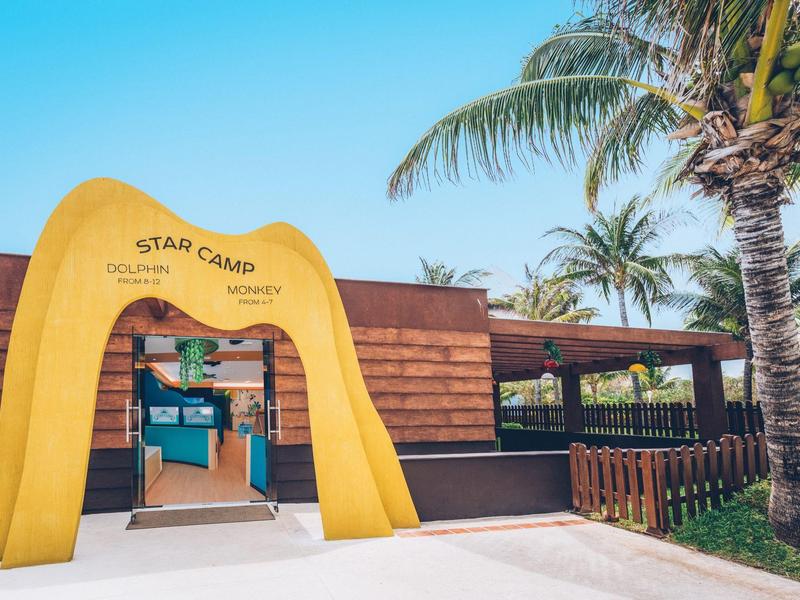 Wejście do obozu na plaży z żółtą arkadą i palmami w tle.