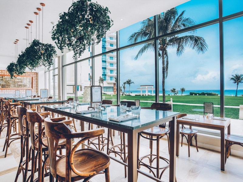 Nowoczesna restauracja z wysokim sufitem, roślinami i widokiem na palmy i morze przez duże okna.