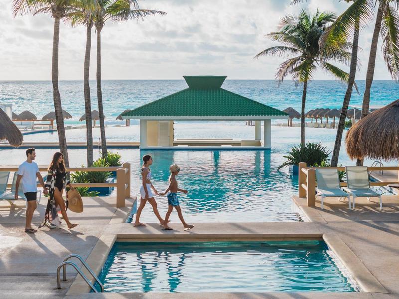 Widok na hotelowy basen z oceanem w tle i spacerującymi gośćmi.
