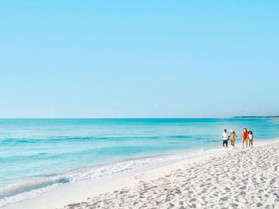 Tre persone che camminano lungo la spiaggia di sabbia bianca con cielo azzurro e mare calmo.