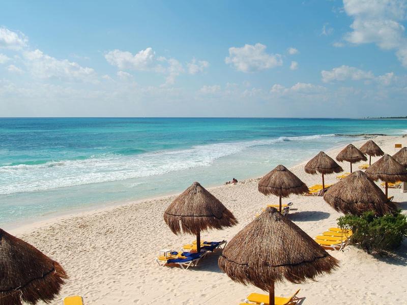 Spiaggia con ombrelloni di palma, sabbia bianca e mare turchese calmo sotto un cielo azzurro.