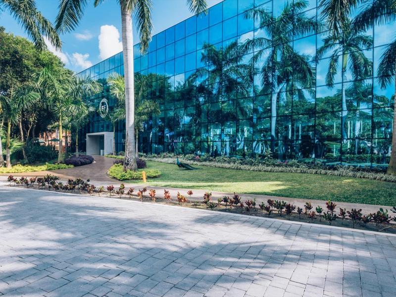 Un'ala di edificio moderno in vetro con palme e fiori in un'ambientazione alberghiera soleggiata.