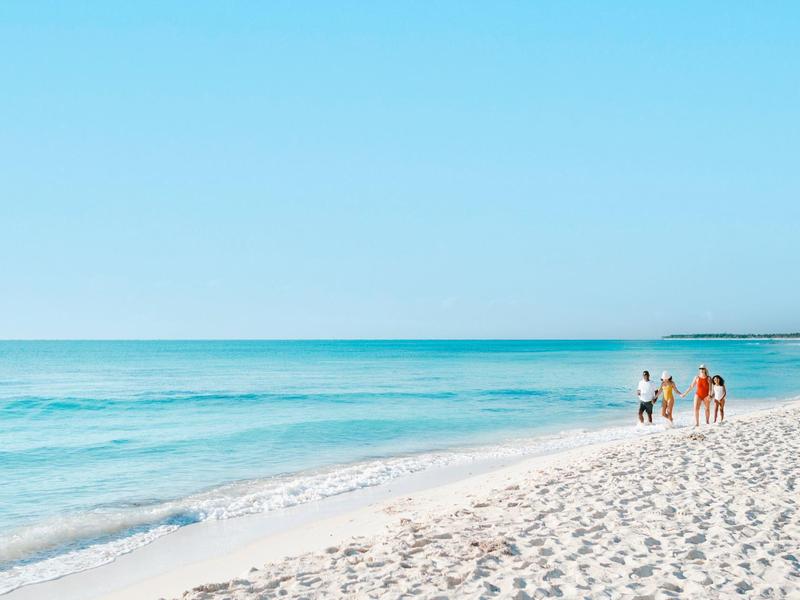 Tre persone che camminano lungo la spiaggia di sabbia bianca con cielo azzurro e mare calmo.