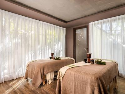 Una stanza luminosa con due lettini da massaggio davanti a grandi finestre con tende bianche.