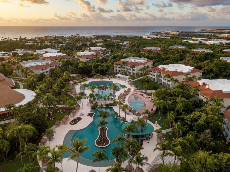 Veduta aerea di un grande resort con piscine circondato da vegetazione rigogliosa e edifici sotto un cielo serale.