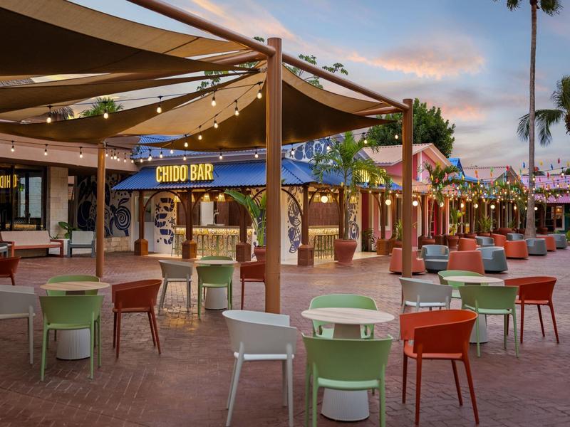 Sedie e tavoli colorati davanti a un bar sotto luci a stringa e palme al tramonto.