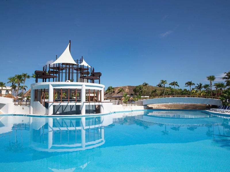 Piscina d'hotel moderna con padiglione e ponte sotto cielo azzurro e palme.