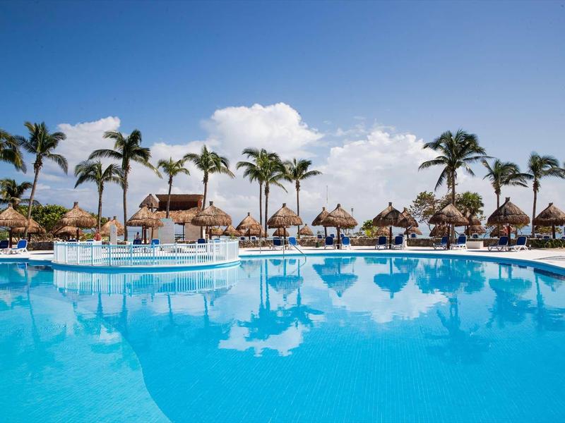 Grande piscina dell'hotel con lettini e palme sotto un cielo azzurro limpido.